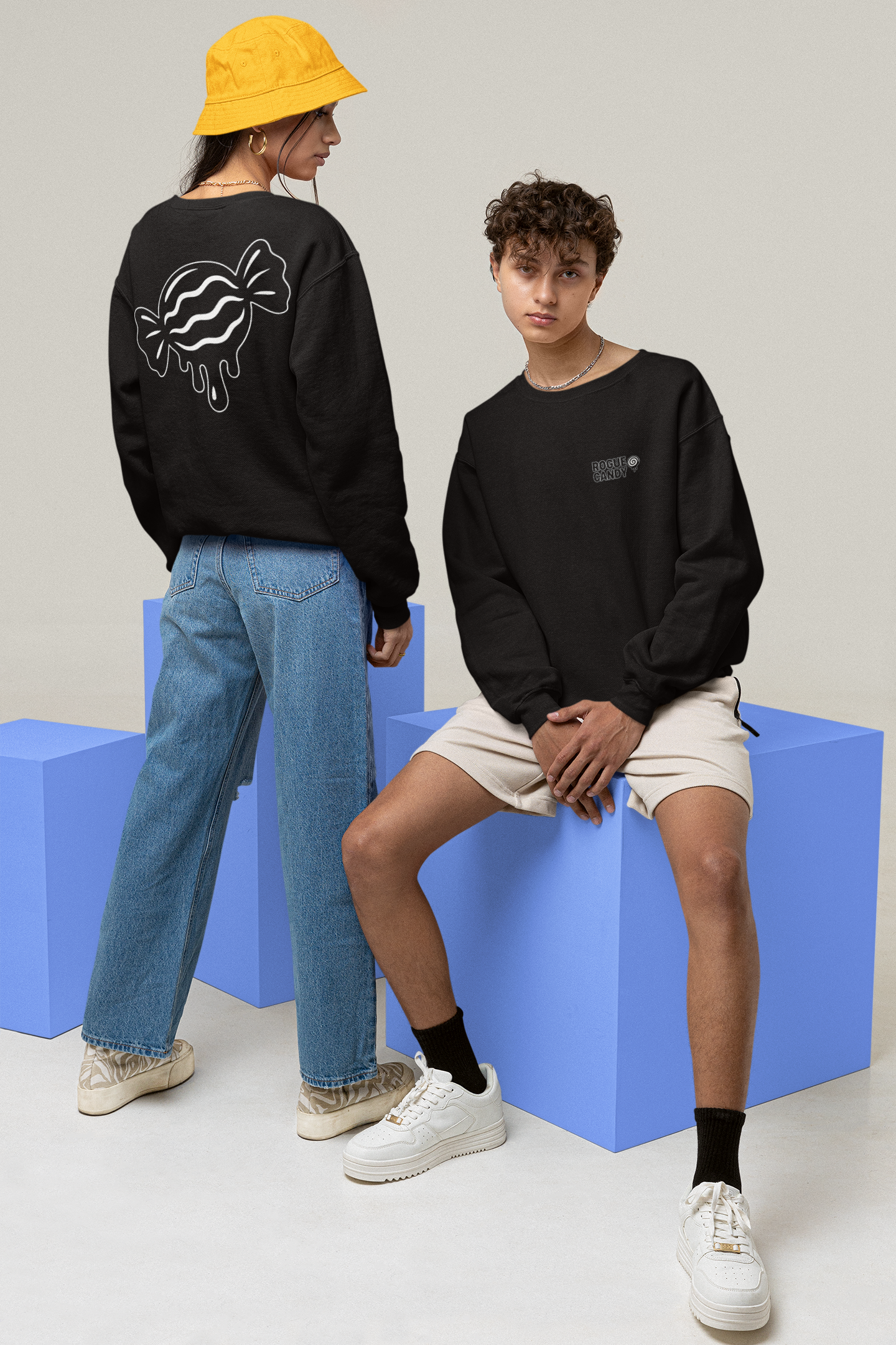 Two people wearing black sweatshirts with different designs, standing and sitting on blue blocks against a light gray background.